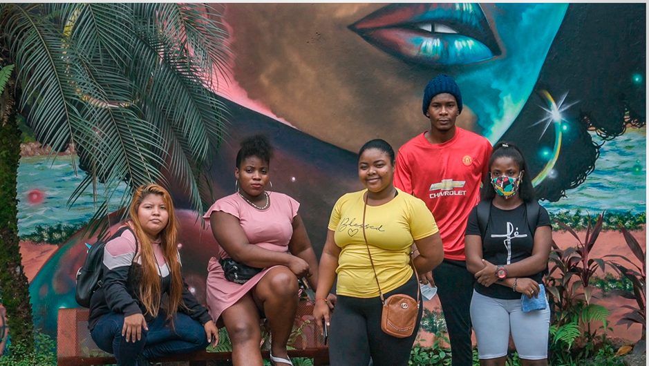 5 Young people of colour in Colombia stand in front of a large and colourful mural of a black woman's head and shoulders.